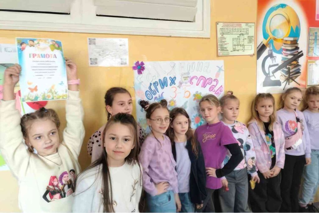 Дівчата з початкових класів біля стенду з грамотами, одна учениця підняла грамоту вгору