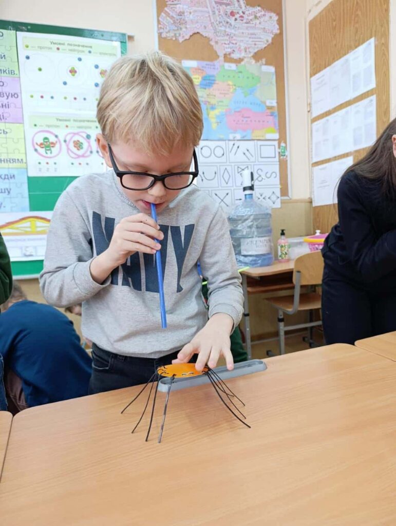 Хлопчик у окулярах вивчає рух паперового павука через соломинку