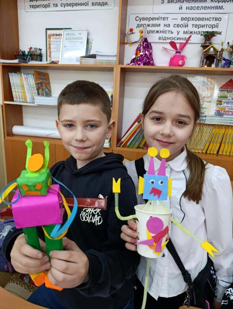 Двоє учнів тримають великих роботів, виготовлених з коробок та чашок